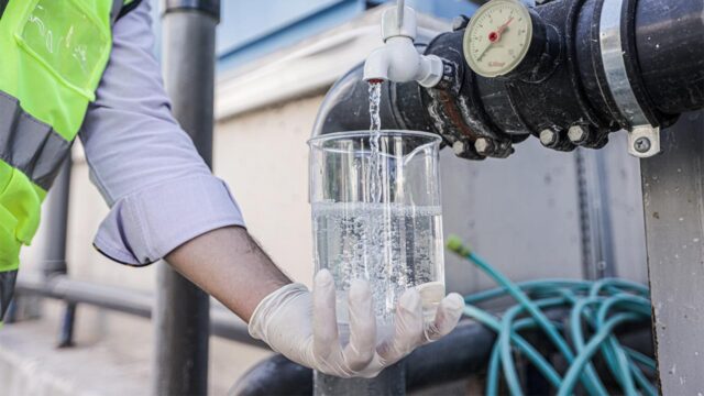 Closeup shot of a person gathering water in a clear glass container from a pipe with a water meter