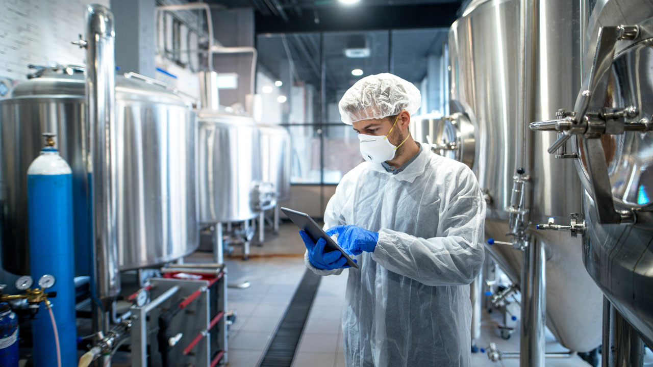 Technician in protective coveralls, gloves, hairnet, and mask using a tablet beside stainless-steel fermentation tanks