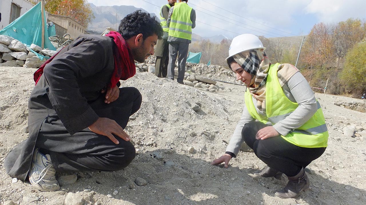 Two people kneel looking down at the ground with a notebook beside them