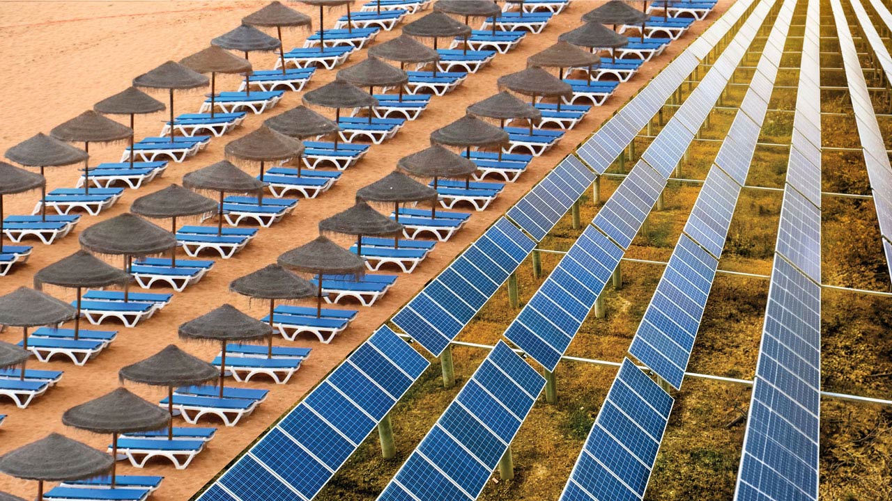 Photograph showing parallel rows of blue solar panels installed on grassy ground adjacent to rows of blue lounge chairs with straw umbrellas on sandy beach