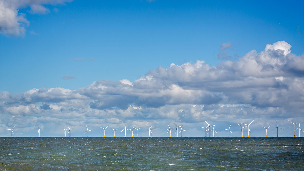 Offshore wind turbines on the horizon with sea in the foreground and a blue sky with a bank of clouds