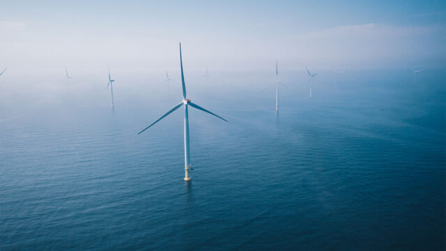 Aerial view of an offshore wind turbine surrounded by sea, with more turbines visible in the distance through mist