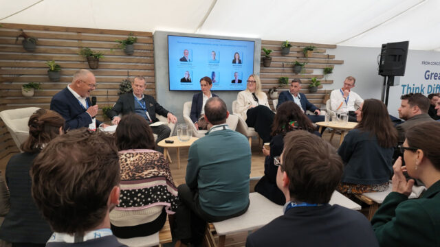 Six people sit on a raised dias with a presentation screen behind and seated audience members in the foreground