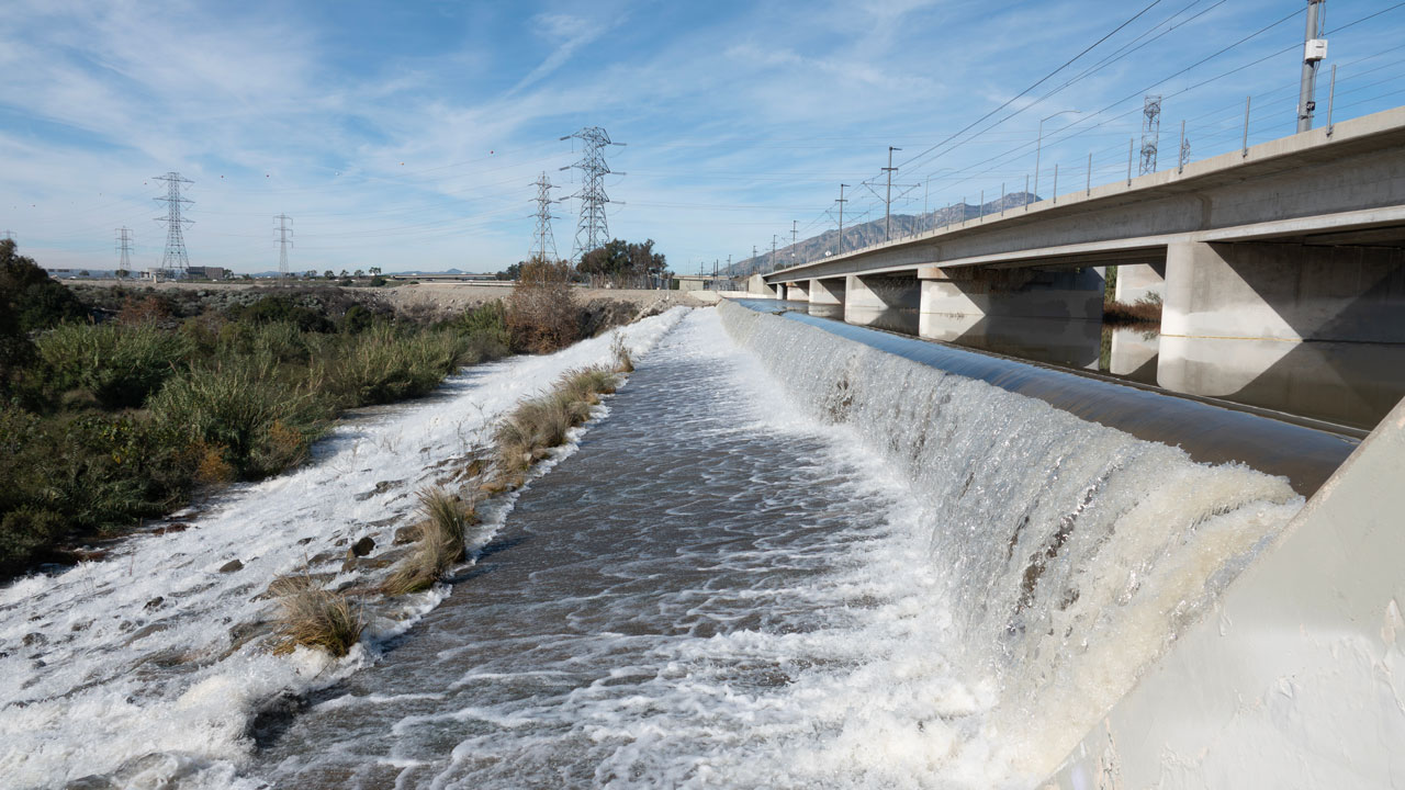 Water rushing through a concrete channel beside a highway bridge under a blue sky