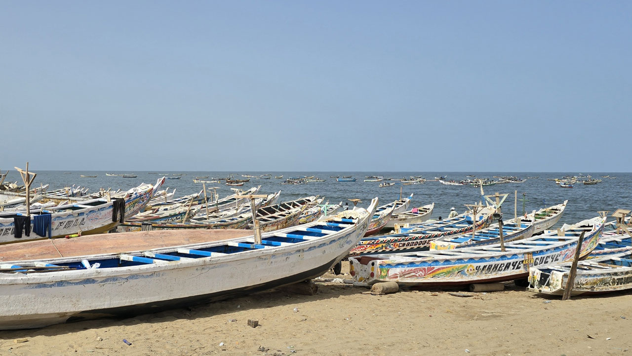 Rows of colourful painted fishing boats lined up on a beach with more fishing boats moored on the sea in background