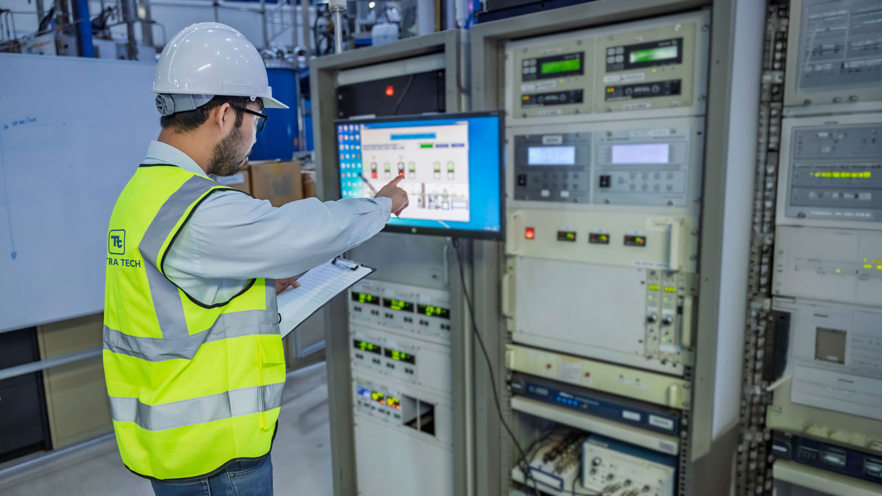 An engineer in a hard hat and safety vest points at a control screen in an equipment room