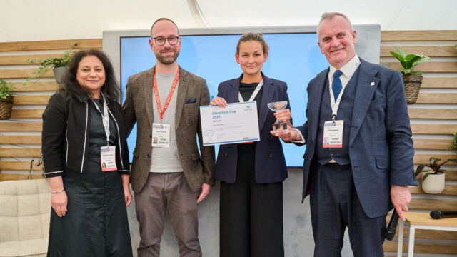 Four people stand on a stage with a presentation screen behind. The two people in the middle are being presented with an award cup and a certificate