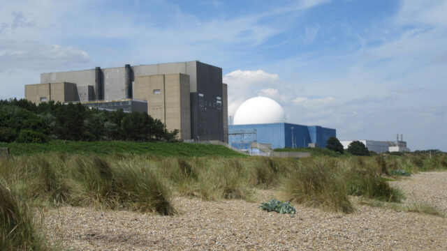 Several industrial buildings against a blue sky with a shingle beach in the foreground