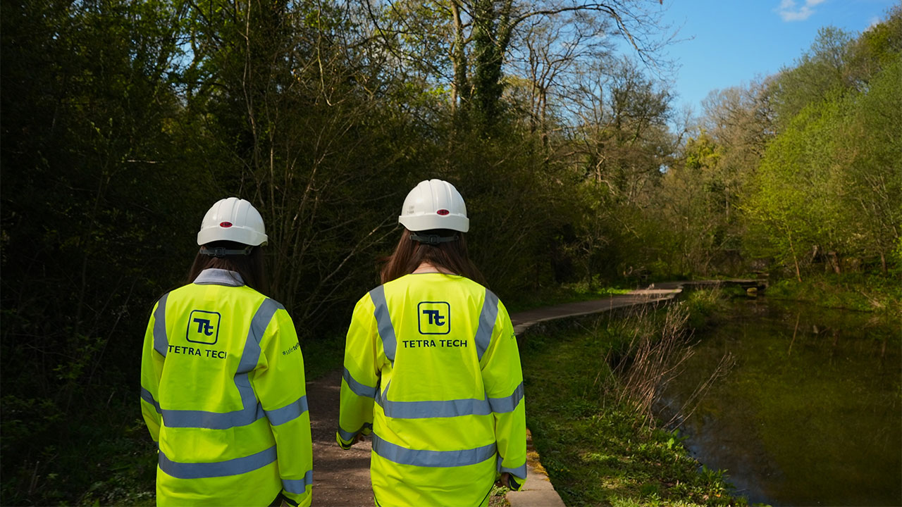 Photograph showing two people wearing bright yellow high-visibility jackets and white helmets walking along a wooded path next to a calm body of water