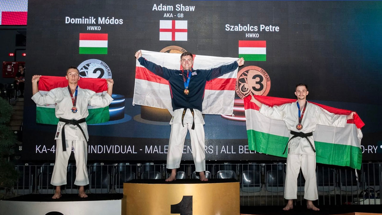Adam Shaw stands atop the #1 podium, holding the English flag, to celebrate his new title as 2026 World Champion, flanked by second and third place competitors