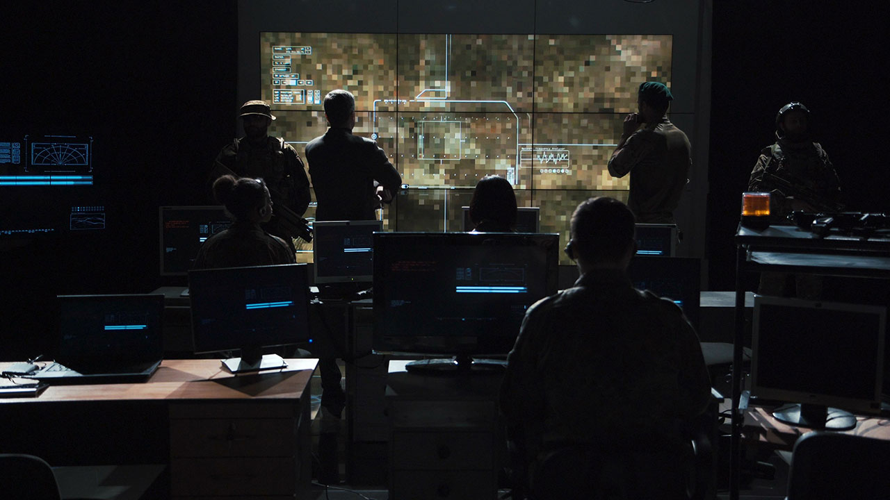 Image of a dark room with the backs of several people looking at a large screen on a wall