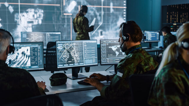 Group of people sitting in a control room at desks with computers and a large digital screen on the wall