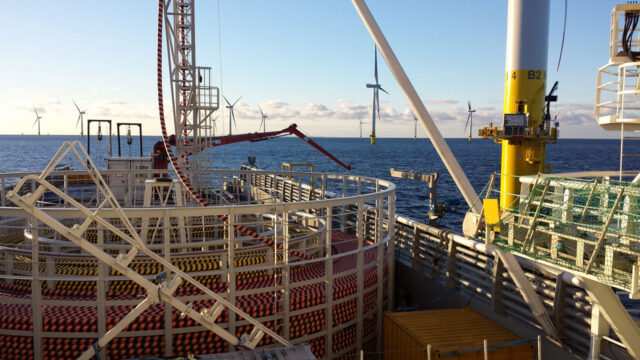 A large circular steel frame containing heavy duty undersea cabling alongside cable-laying equipment in the foreground and offshore wind turbines in the background