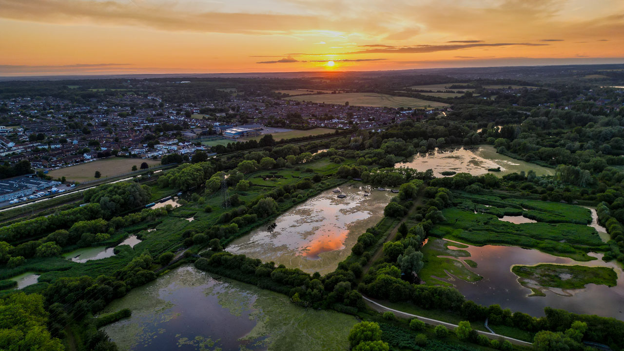Aerial view of rural landscape at sunset showing flooded fields in the foreground with a river and a housing estate in the background