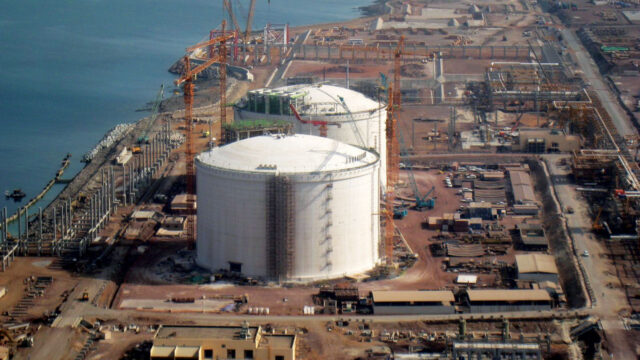 Aerial view of energy facility under construction with large storage tanks, cranes, piping, and waterfront infrastructure